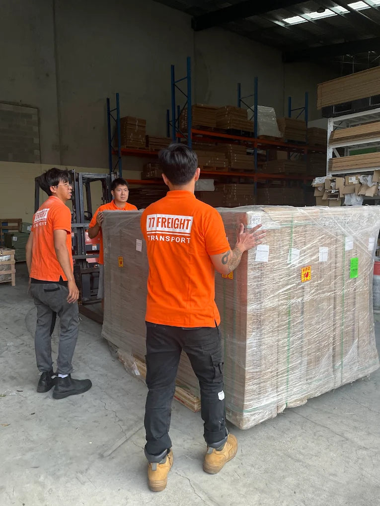 Three workers in orange '77 Freight Transport' shirts moving a large pallet of boxes in a warehouse.